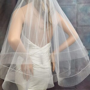 May include: A white single-tier bridal veil with a beaded trim and two layers of tulle. The veil is shown on a mannequin wearing a white dress.