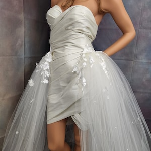 May include: A white strapless wedding dress with a fitted bodice and a flowing tulle skirt. The skirt has a high-low design, with a shorter front and a longer train in the back. The dress is decorated with delicate lace appliques.