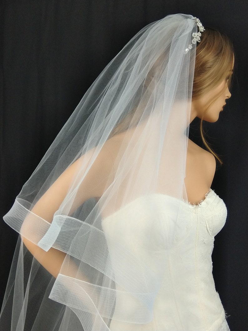 May include: A white tulle wedding veil with a single-tier design and a beaded headband. The veil has a scalloped edge and a satin ribbon trim.