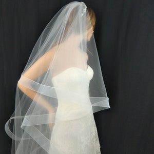 May include: A white bridal veil with a single layer of tulle and a delicate beaded trim. The veil is draped over a white wedding dress.
