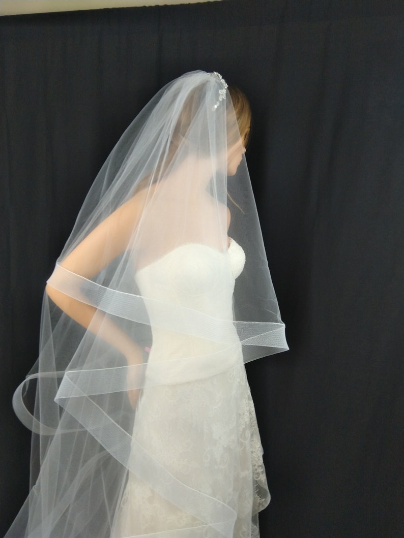 May include: A white bridal veil with a single layer of tulle and a delicate beaded trim. The veil is draped over a white wedding dress.