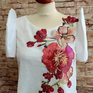 May include: White top with short sleeves and a floral embroidery design. The embroidery features pink, red, and yellow flowers.