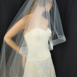 May include: A white bridal veil with a single layer of tulle and a scalloped edge. The veil is worn over a white strapless wedding dress with lace detailing.