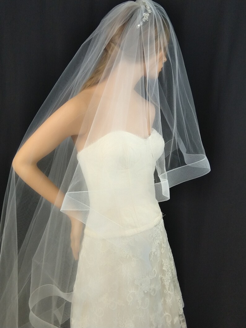 May include: A white bridal veil with a single layer of tulle and a scalloped edge. The veil is worn over a white strapless wedding dress with lace detailing.