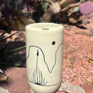May include: A cylindrical ceramic tumbler with a cream-colored glaze. The tumbler features a black line drawing of a desert landscape with a sun and a plant. The tumbler is approximately 4 inches tall.