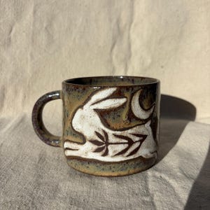 May include: A ceramic mug with a speckled brown and green glaze featuring a white relief carving of a rabbit leaping toward a crescent moon. The rabbit's body is decorated with a dark brown leaf pattern, and the mug has a sturdy side handle.
