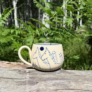 May include: Cream-colored ceramic mug with a blue interior and a curved handle. The mug features a blue line drawing of horses and a blue dot. The mug is sitting on a wooden surface with green foliage in the background.