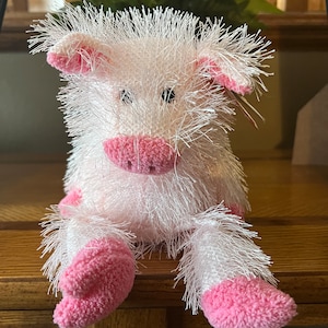 May include: A plush pig toy with white, fluffy fur and pink accents on the snout, ears, and feet. The stuffed animal is sitting on a wooden surface. The pig has black button eyes.