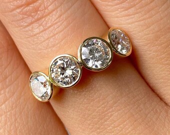May include: A gold ring featuring four round, brilliant-cut clear stones set in a bezel style. The stones are arranged in a horizontal row across the band, with the center two stones being larger than the two stones on the outer ends.