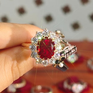 Puede incluir: Un anillo de oro con una gran piedra roja ovalada rodeada de piedras transparentes más pequeñas. El anillo se sostiene en una mano, y hay otros anillos en el fondo.