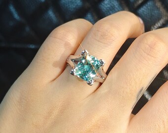 May include: A silver ring with a large, square, light blue gemstone is displayed on a hand. The ring has a unique setting with smaller accent stones. The background is a black, quilted texture. The Leyloon Jewelry logo is visible.