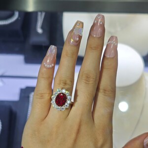 Könnte beinhalten: Ein goldener Ring mit einem großen, oval geschliffenen, tiefroten Edelstein, umgeben von kleineren, klaren Edelsteinen. Der Ring befindet sich an einer Hand mit gepflegten Nägeln.