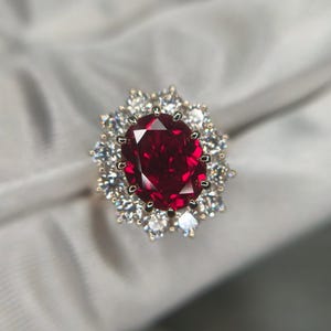 May include: A gold ring featuring a large, oval-cut, deep red gemstone surrounded by smaller, clear gemstones. The ring is set against a soft, white background. The ring is a piece of jewelry.