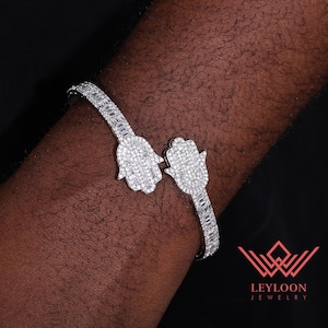 Puede incluir: Una pulsera abierta de tono plateado con dos motivos de mano de Hamsa en los extremos. Toda la superficie de la pulsera y los dijes de Hamsa están incrustados con numerosas piedras pequeñas, claras y brillantes que captan la luz.