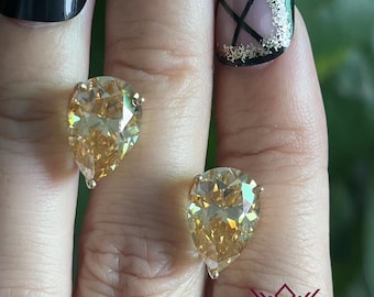 May include: A pair of pear-shaped earrings with a gold setting and a champagne-colored gemstone. The earrings are shown on a hand with black nail polish.