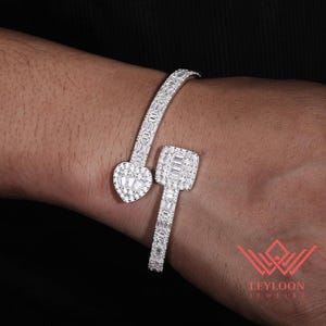 Puede incluir: Una pulsera abierta de tono plateado con un terminal en forma de corazón y otro cuadrado, ambos incrustados con numerosas piedras transparentes pequeñas de corte baguette y redondo. La banda también está pavimentada con piedras.