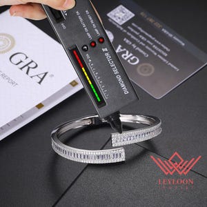 Puede incluir: Una pulsera abierta de tono plateado con una hilera de piedras claras en corte baguette bordeadas por pequeñas piedras redondas. Un dispositivo de prueba Diamond Selector II negro está sobre la pulsera mostrando luces verdes.