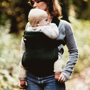 Pu&ograve; includere: Una donna con un maglione grigio porta un bambino in un marsupio verde scuro. Il bambino indossa un maglione bianco e ha i capelli biondi. La donna e il bambino sono in un ambiente boschivo.