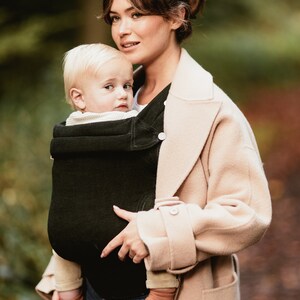 Pu&ograve; includere: Una donna con un cappotto beige porta un bambino in un marsupio verde scuro. Il bambino indossa una camicia bianca e calzini marroni.