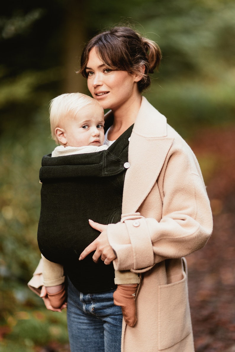 Pu&ograve; includere: Una donna con un cappotto beige porta un bambino in un marsupio verde scuro. Il bambino indossa una camicia bianca e calzini marroni.