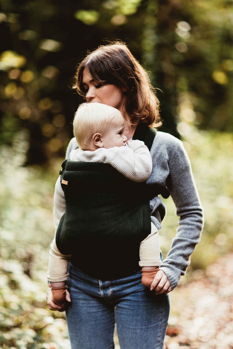 Pu&ograve; includere: Una donna con un maglione grigio porta un bambino in un marsupio verde scuro. Il bambino indossa un maglione bianco e ha i capelli biondi. La donna e il bambino sono in un ambiente boschivo.