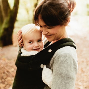Pu&ograve; includere: Una donna con un maglione grigio porta un bambino in un marsupio verde scuro. Il bambino guarda la donna e sorride.