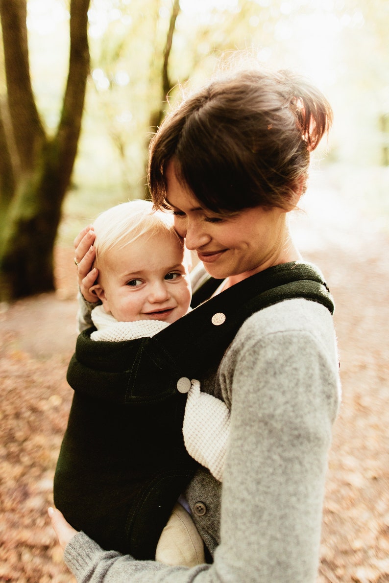 Pu&ograve; includere: Una donna con un maglione grigio porta un bambino in un marsupio verde scuro. Il bambino guarda la donna e sorride.