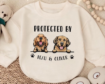 May include: Cream-colored baby sweatshirt with the text "PROTECTED BY" above illustrations of two golden retrievers. Below the dogs are the words "BEAU & OLIVER" and paw prints. The sweatshirt is on a woven surface with a stuffed animal and baby shoes.