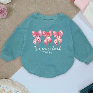 May include: Teal-colored baby sweatshirt with three watercolor bows in pink and red hues. The bows have heart and polka dot patterns. The text "You are so loved" and "JOHN 3:16" are printed below the bows.