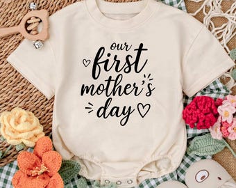May include: A cream-colored baby onesie with short sleeves and a black graphic that reads "our first mother's day" with a heart symbol on either side of the text.