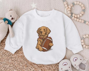 May include: White long-sleeved baby bodysuit featuring a cartoon golden retriever holding a brown football. The dog has a red collar. The bodysuit is surrounded by a teddy bear, baby shoes, and a wooden bead garland.