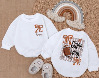 May include: Two white baby rompers with football-themed designs. One has a bow and the name "Emma", the other features a football, the words "Game day", and decorative bows. Two pairs of beige baby shoes are also visible.