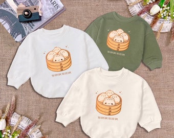 May include: Three baby sweatshirts in white, cream, and olive green. Each features a cartoon illustration of dim sum in a bamboo steamer with the text "YOU DIM SUM, YOU LOVE SUM."