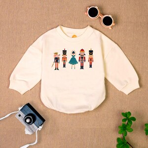 May include: A white long-sleeved sweatshirt with a graphic of a ballerina and nutcracker characters. The sweatshirt has a crew neck and is made of a soft, comfortable fabric.