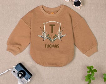 May include: A brown long-sleeved sweatshirt with a graphic of a crest with the letter "T" inside and two ducks flying above. The text "THOMAS" is printed below the crest.