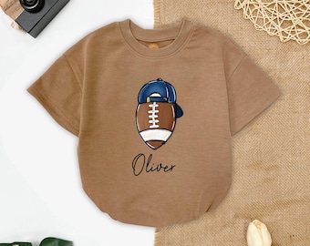 May include: A short-sleeved, brown baby bodysuit with a football graphic wearing a blue baseball cap. The name "Oliver" is written in cursive below the football design. The bodysuit is made of soft material.