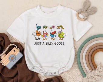 May include: White baby onesie with a graphic of four cartoon geese, each wearing different accessories like a snorkel, sunglasses, and a hat. The text "JUST A SILLY GOOSE" is printed below the geese. A wooden toy camera and a rainbow are in the background.