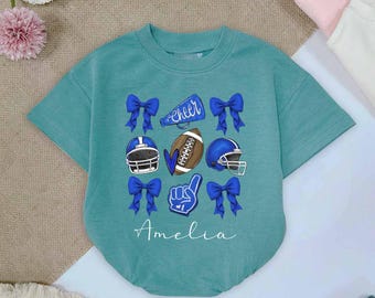 May include: Teal short-sleeved baby bodysuit with a football theme. The design includes blue bows, football helmets, a football, a foam finger, and the word "Cheer". The name "Amelia" is printed at the bottom.