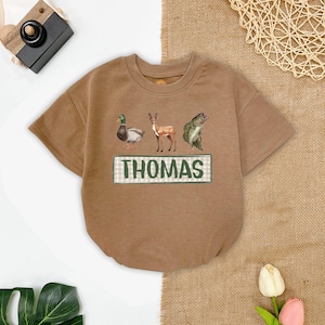 May include: A brown short-sleeved t-shirt with a green and white checkered rectangle that says "THOMAS" in green letters. The shirt has a duck, deer, and fish graphic printed on it.