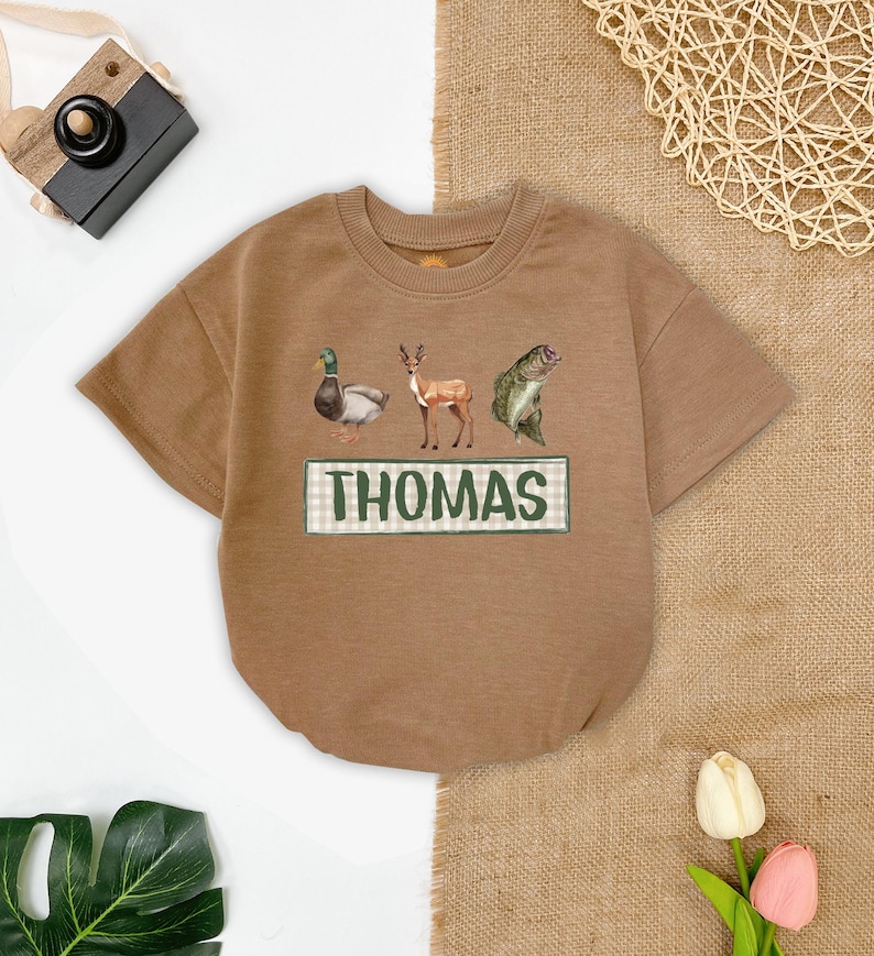 May include: A brown short-sleeved t-shirt with a green and white checkered rectangle that says "THOMAS" in green letters. The shirt has a duck, deer, and fish graphic printed on it.