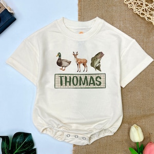 May include: A white baby onesie with a green and brown plaid banner that says "THOMAS" and three images of a duck, a deer, and a fish.