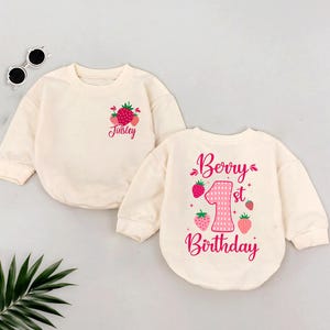 May include: Two cream-colored long-sleeved shirts with pink strawberry designs. One shirt says "Berry 1st Birthday" and the other says "Tinsley".
