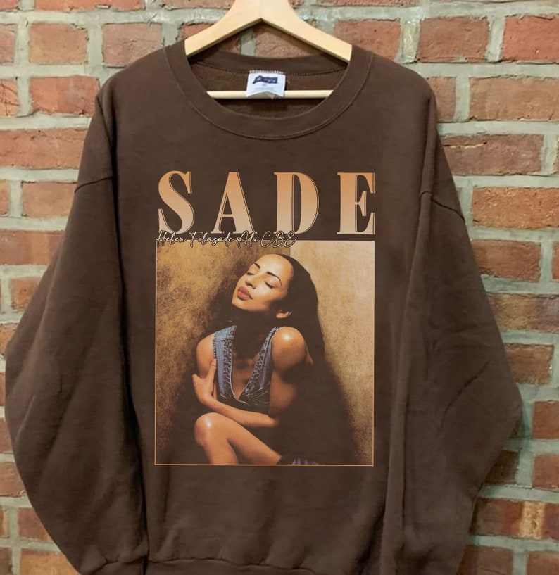 May include: Brown sweatshirt with a vintage graphic print of Sade, the singer, with the text "SADE Helen Folasade Adu CBE" on the front.