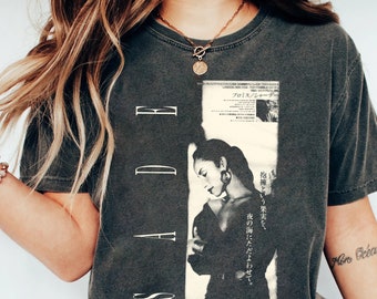 May include: A dark gray t-shirt with a black and white image of a woman with the text "SADE" and Japanese text on the front. The text on the shirt reads "Promise/Sade" and "The fruit of a hug, I let it drift in the night sea."
