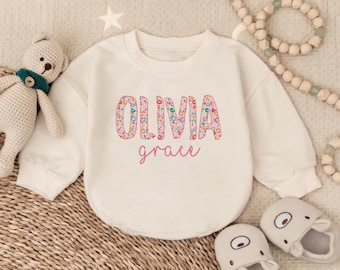 May include: Cream-colored baby sweatshirt with the name "Olivia" in a floral pattern and the name "grace" in pink script. The sweatshirt is paired with matching baby shoes and a stuffed bear.