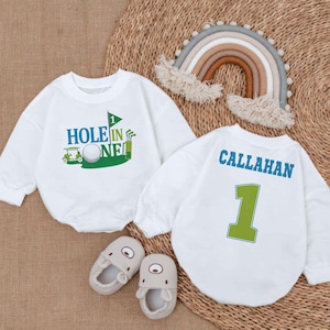 May include: Two white long-sleeve shirts with golf-themed designs. One shirt says "Hole in One" with a golf cart and golf ball. The other shirt says "CALLAHAN" with the number "1" in green.