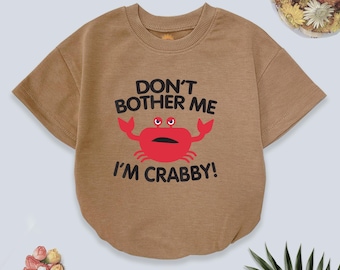 May include: A tan short-sleeved baby bodysuit with the text "DON'T BOTHER ME I'M CRABBY!" and a red crab graphic. The bodysuit is made of soft material and has a round neckline.