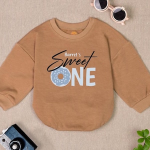 Può includere: Una felpa marrone a maniche lunghe con il testo "Barret's Sweet One" e una grafica di ciambella blu.