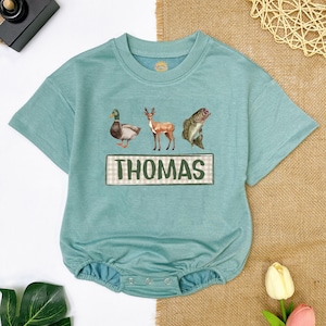 May include: A teal blue baby onesie with a duck, deer, and fish graphic. The onesie has a green and white checkered name tag with the name "THOMAS" printed on it.