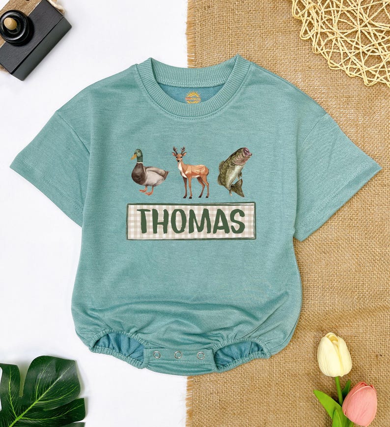 May include: A teal blue baby onesie with a duck, deer, and fish graphic. The onesie has a green and white checkered name tag with the name "THOMAS" printed on it.